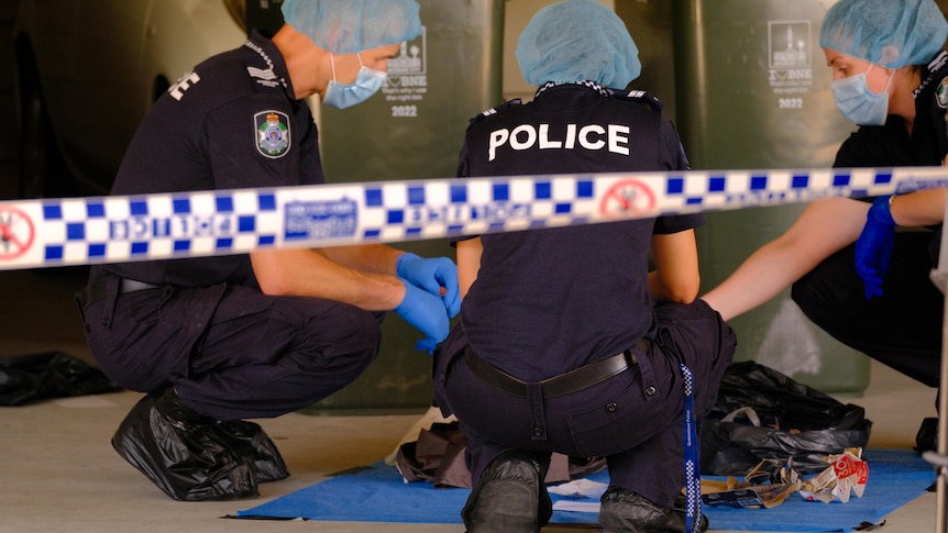 Police crouch over evidence behind crime scene tape.