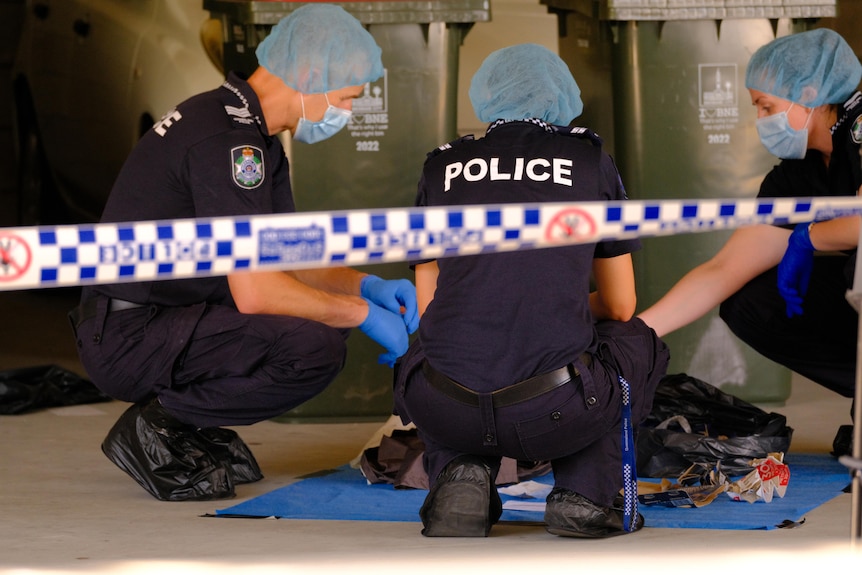 Police crouch over evidence behind crime scene tape.