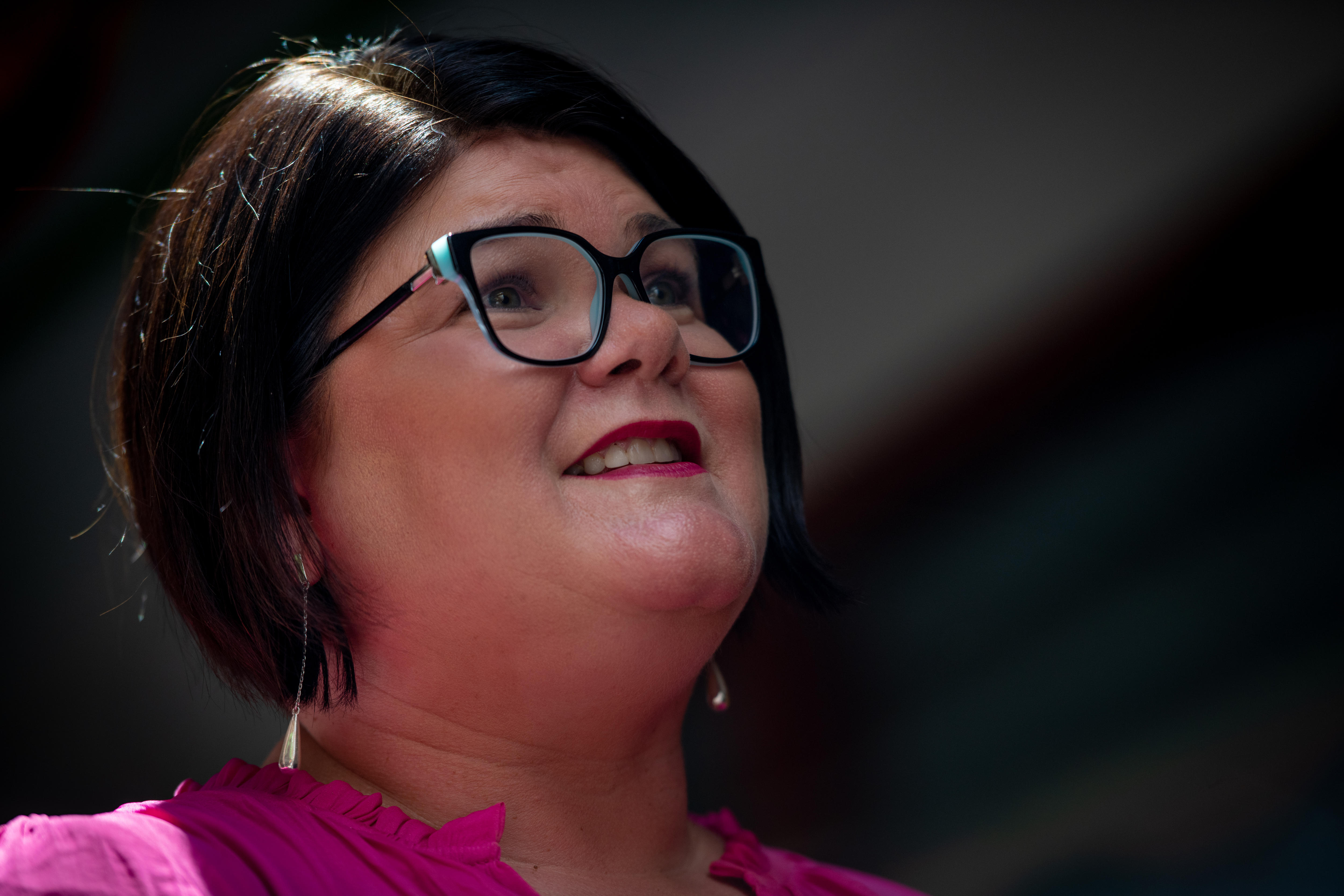 Close de uma mulher com cabelo curto e escuro e óculos quadrados pretos e azul-petróleo, olhando para longe da câmera