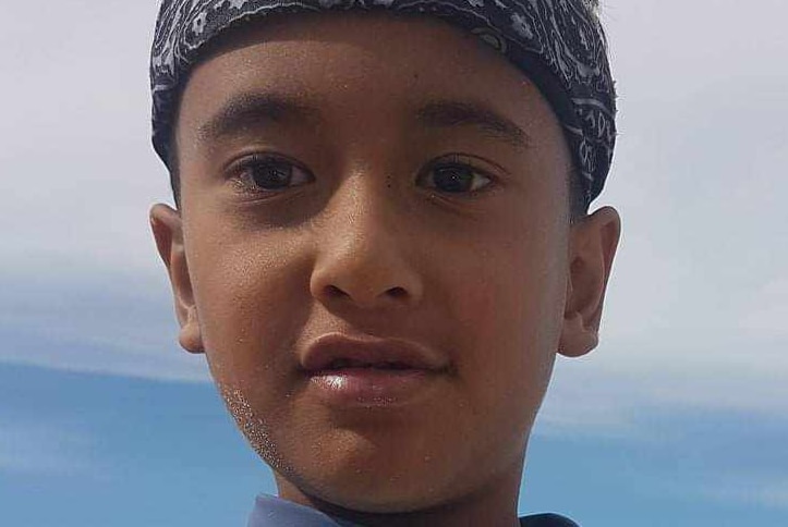 A young boy wears a rash top and bandana at the beach on a bright day with cloud.