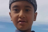 A young boy wears a rash top and bandana at the beach on a bright day with cloud.