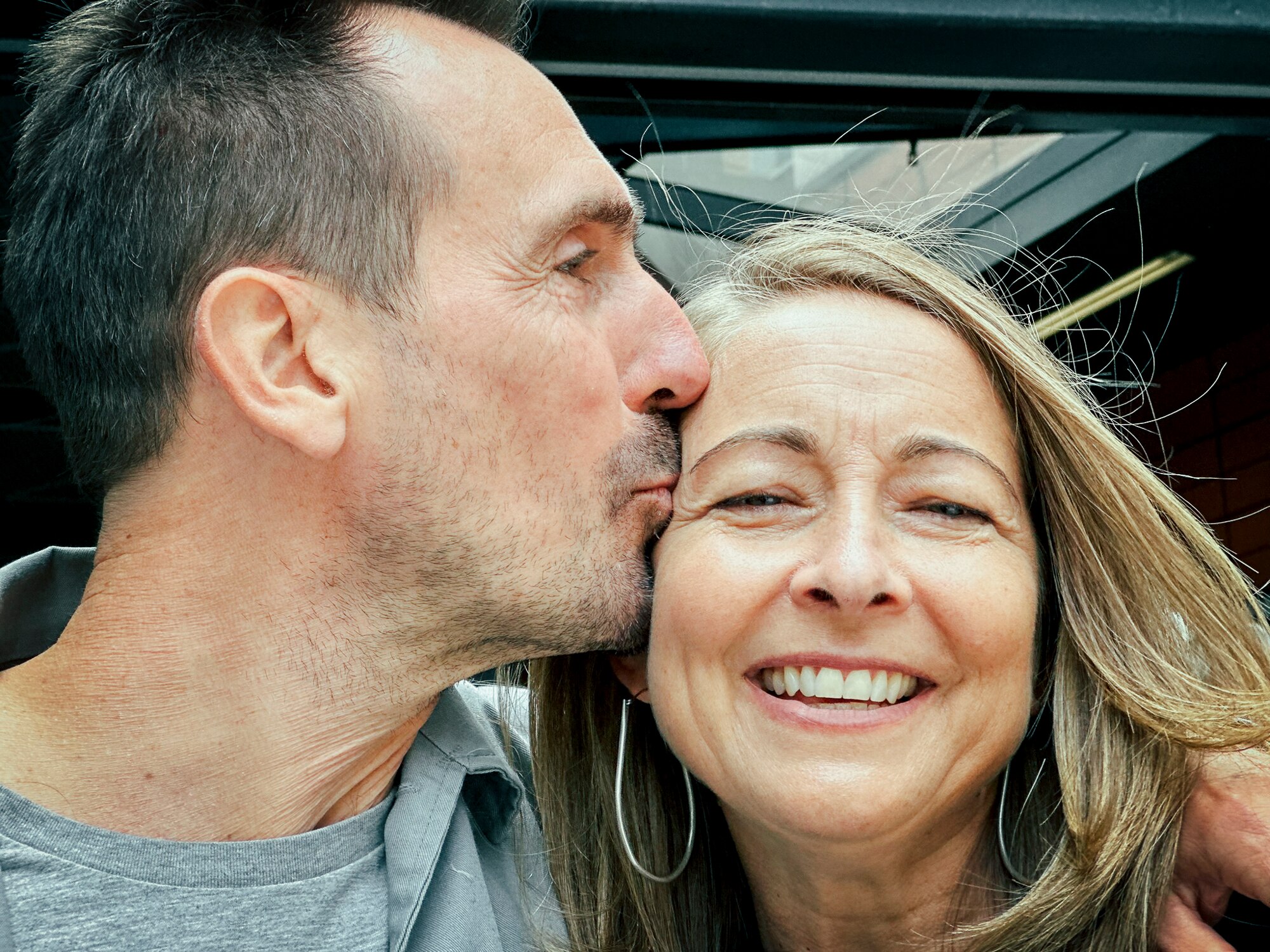 A close up of a man kissing his wife on the side of the face. He is side on and she faces the camera and smiles.