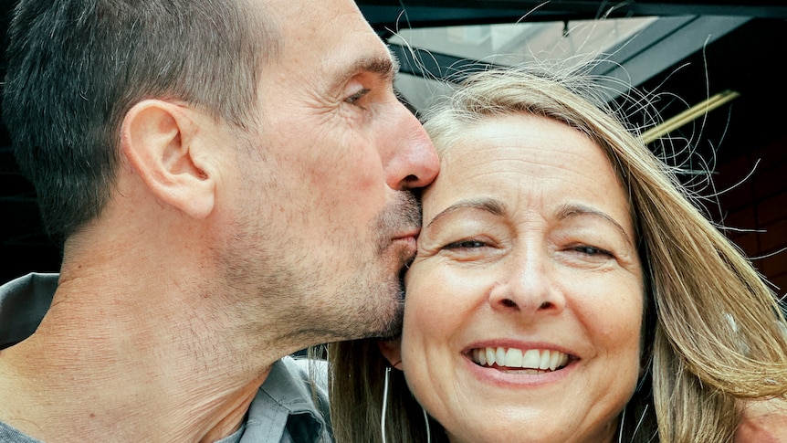 A close up of a man kissing his wife on the side of the face. He is side on and she faces the camera and smiles.