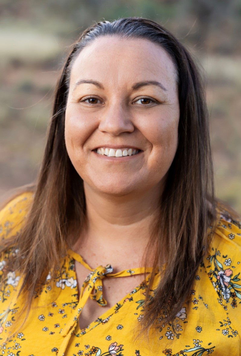 A woman with brown hair wearing a yellow patterned top smiling.