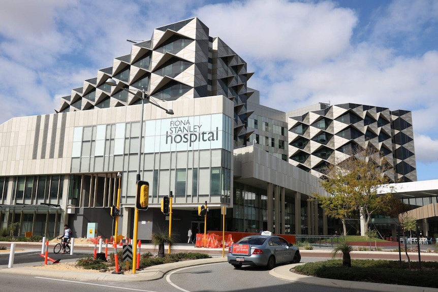 A taxi approaches the entrance to Fiona Stanley Hospital in Perth as a cyclist prepares to cross the crosswalk.