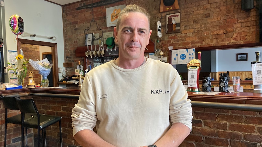 A picture of a man in a white jumper standing in front of a bar.