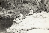 Wide black-and-white archive shot of a group of naked women sitting on a large rock next to a swimming hole.
