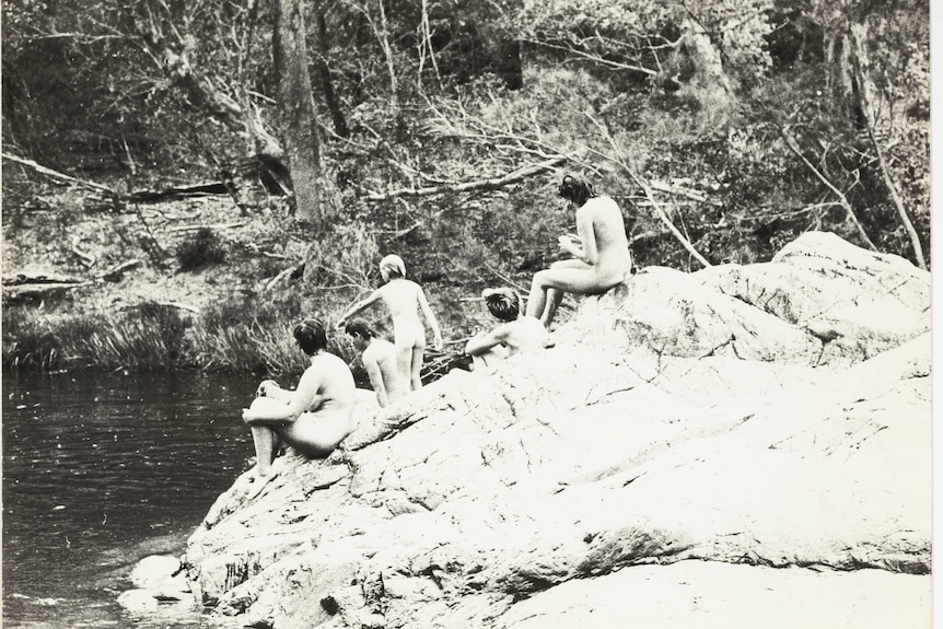 Wide black-and-white archive shot of a group of naked women sitting on a large rock next to a swimming hole.