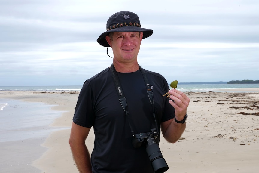 Dion holds a small green plant on a beach with his camera around his neck.