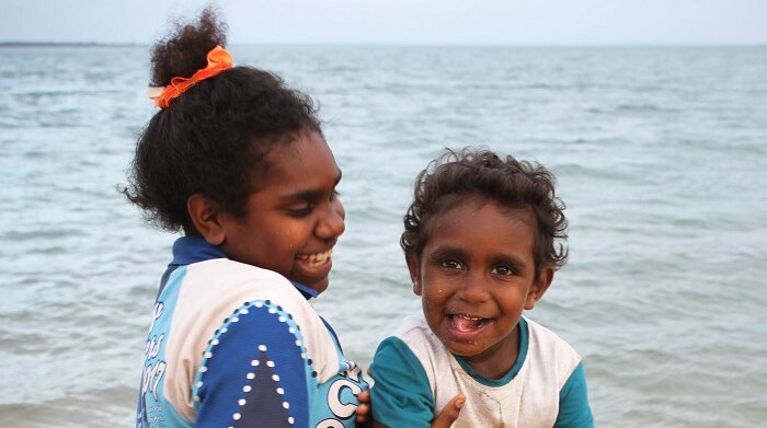 A photo of a young Indigenous girl with a small Indigenous boy on her lap laughing while they fish on the beach at Cullen Point.