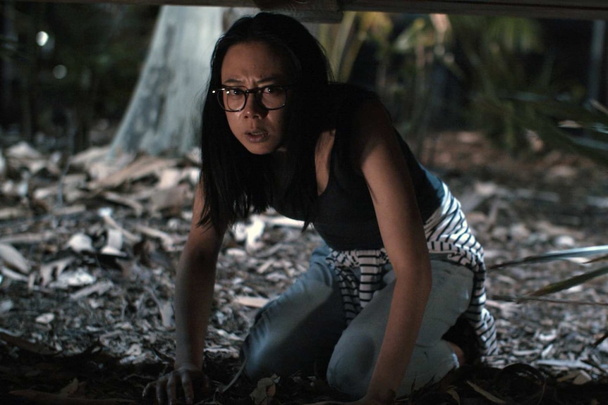 Young woman with long black hair, wearing glasses and a dark tee and jeans, crouches on leaf-covered ground outdoors, at night.