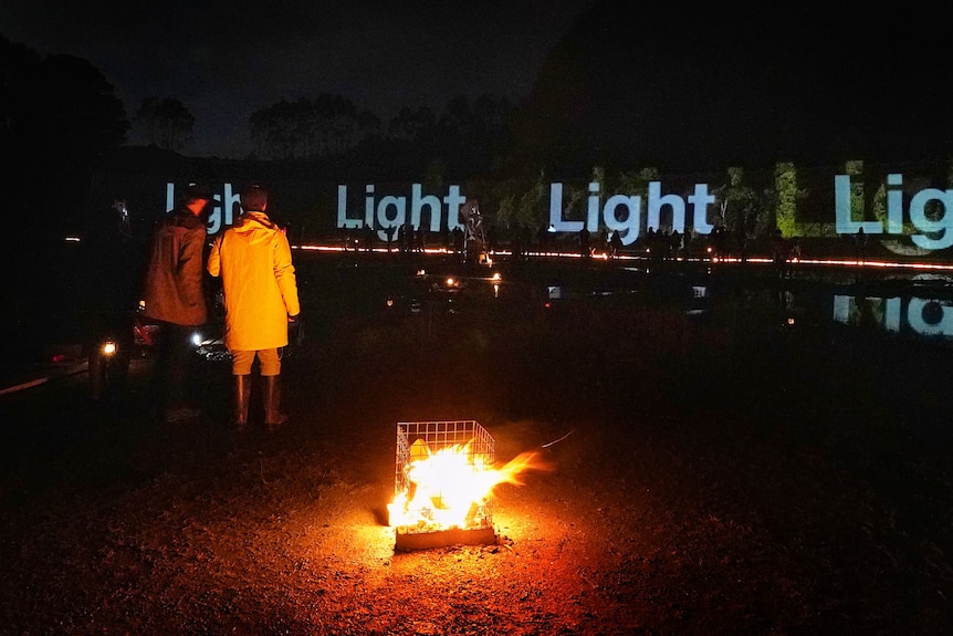 Two people wearing bright raincoats stand in front of a small fire in a drum at night. Behind the word "light" is projected.