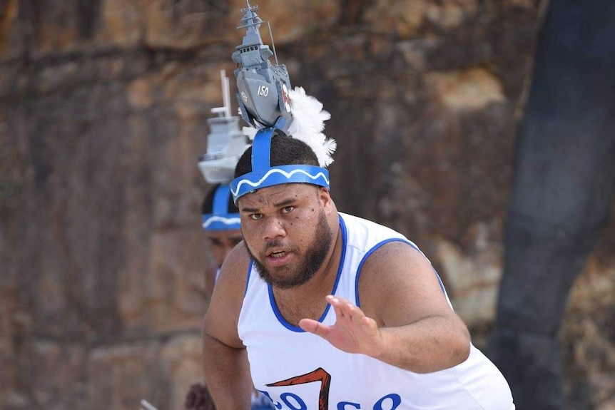 Young Torres Strait Islander man in traditional dress dancing on the sand with rocks in background.
