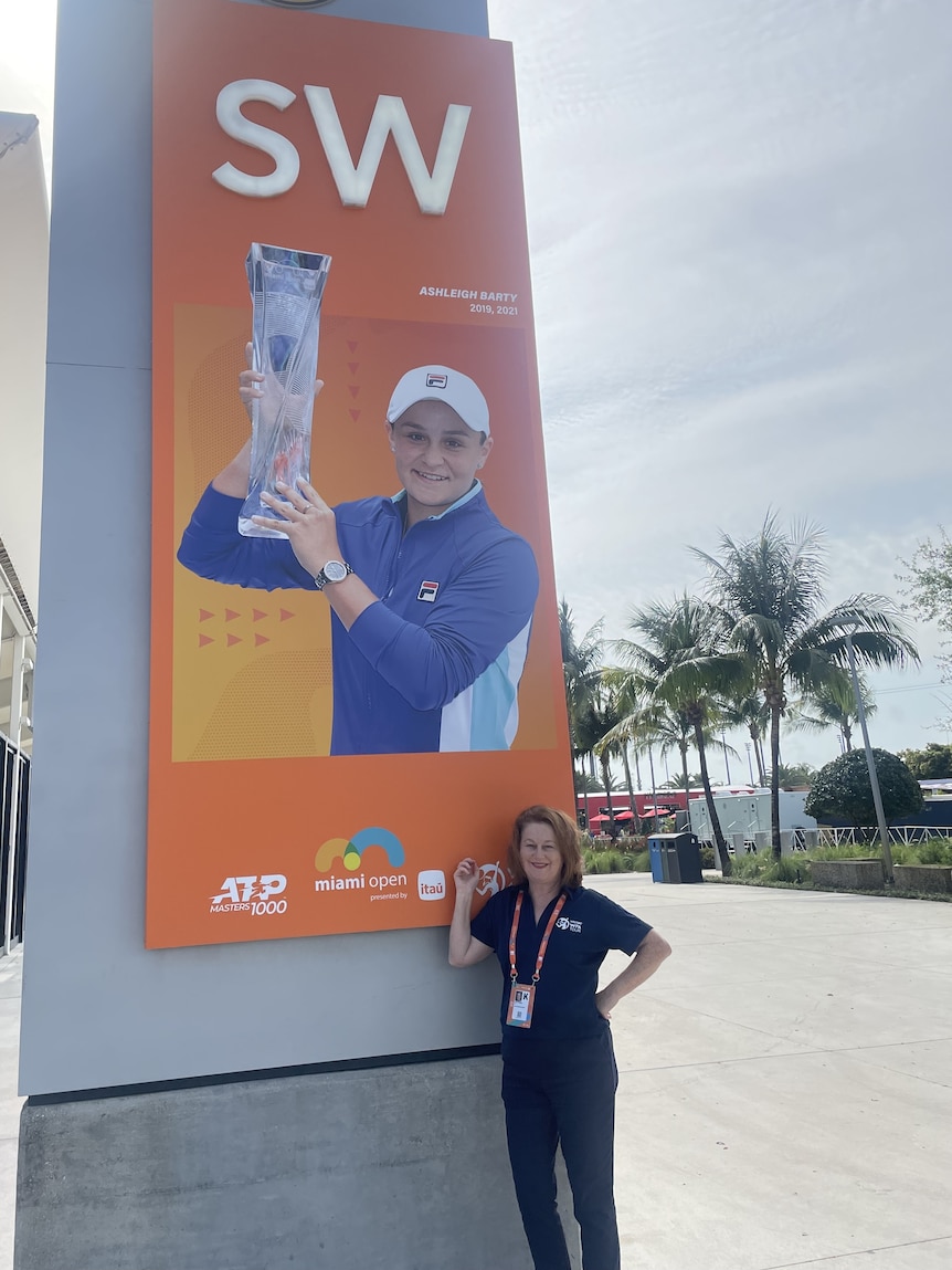 Deirde stands next to a large sign with Ash Barty holding a trophy, she is pointing at the sign in a blue polo shirt.
