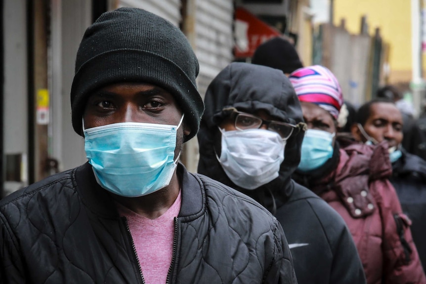 A line of African American people stand in Harlem wearing medical face masks and beanies.