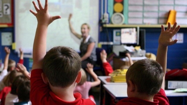 Two boys sitting in a classrooms with one hand raised, and the teacher in the background.