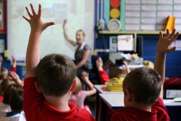 两个男孩坐在教室里，举起一只手，老师在后面。