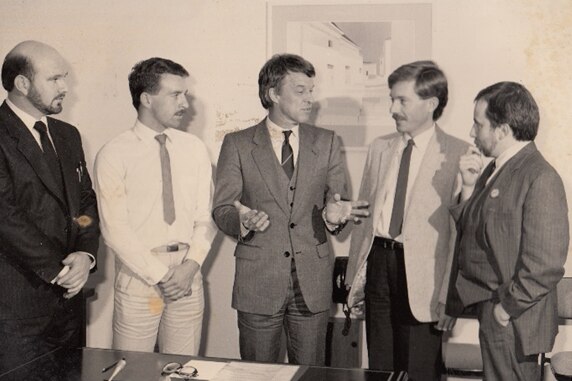 Five men wearing suits and ties speak inside an office; the man in the centre gestures with his hands.
