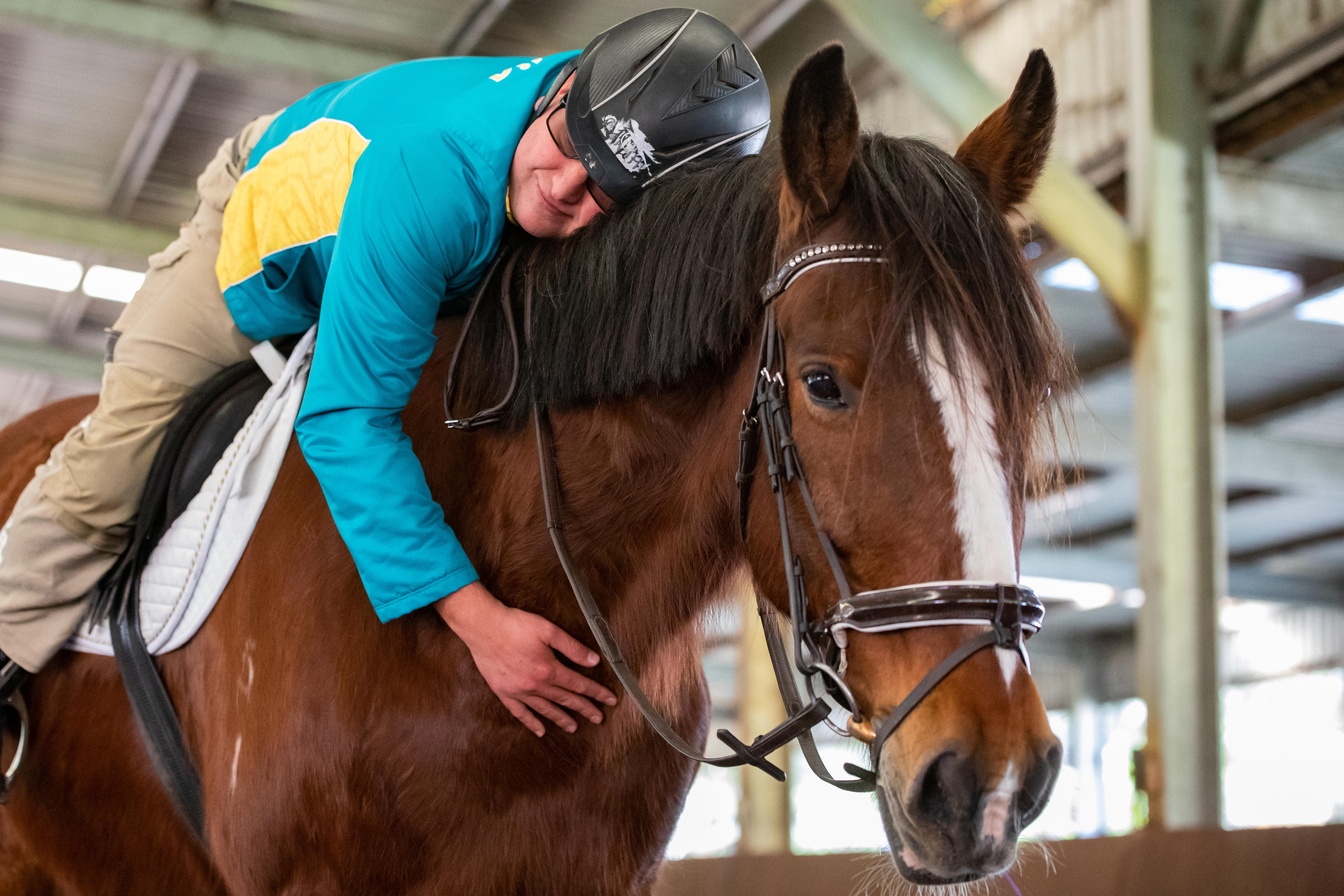 A man wearing a shirt with Australian sits on top of a horse while hugging it