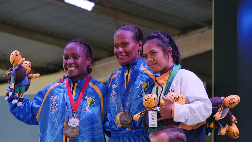 Three smiling people stand on a podium while wearing medals.