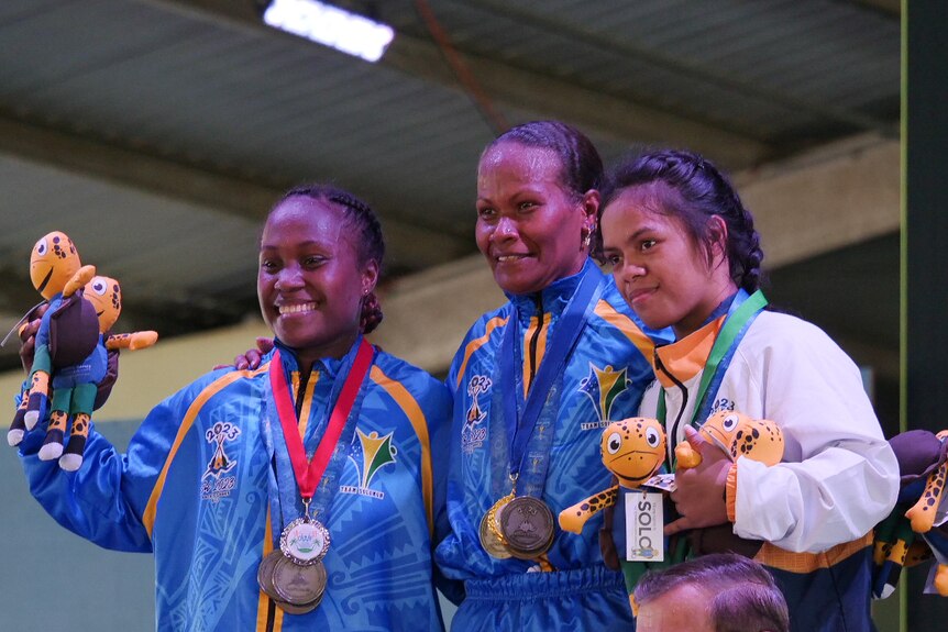 Three smiling people stand on a podium while wearing medals.