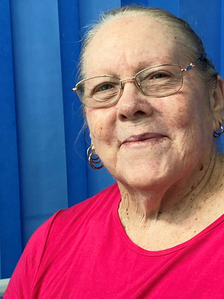 A 75-year-old woman wearing glasses and gold earrings smiles for a photo in front of a blue curtain.