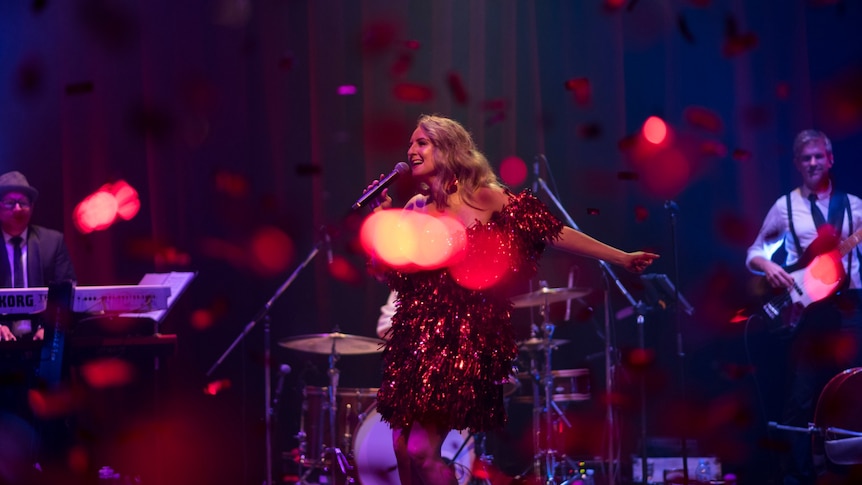 Naomi Price sings on stage in a red tinsel dress as red confetti rains down.