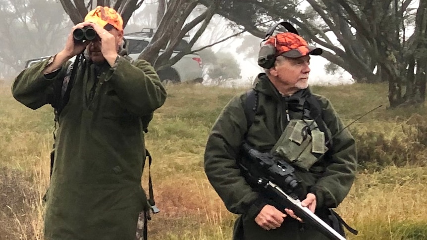 Two men stand in a field dressed in hunting garb. One holds binoculars to his eyes, the other holds a gun.