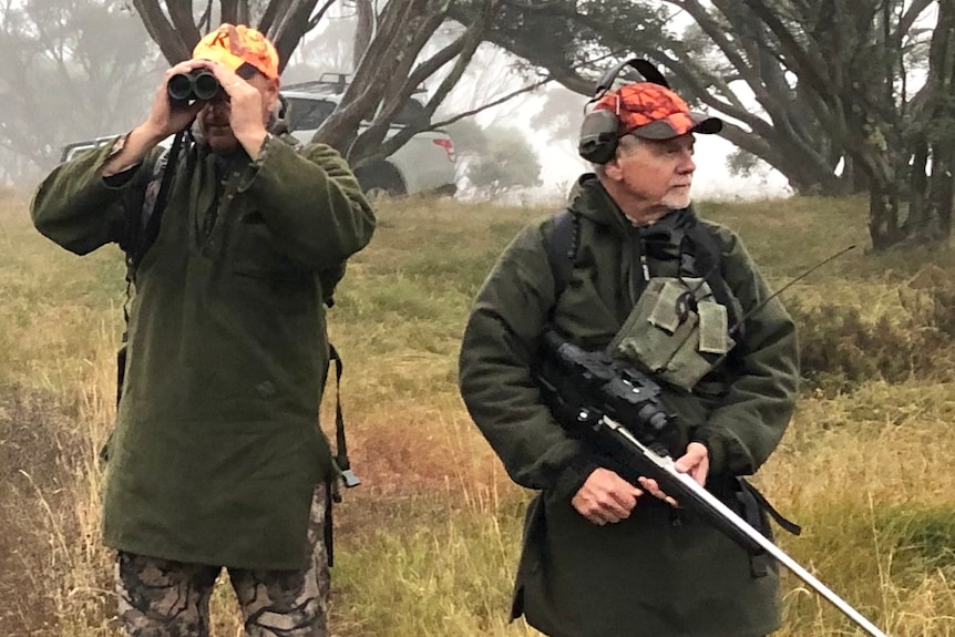 Two men stand in a field dressed in hunting garb. One holds binoculars to his eyes, the other holds a gun.