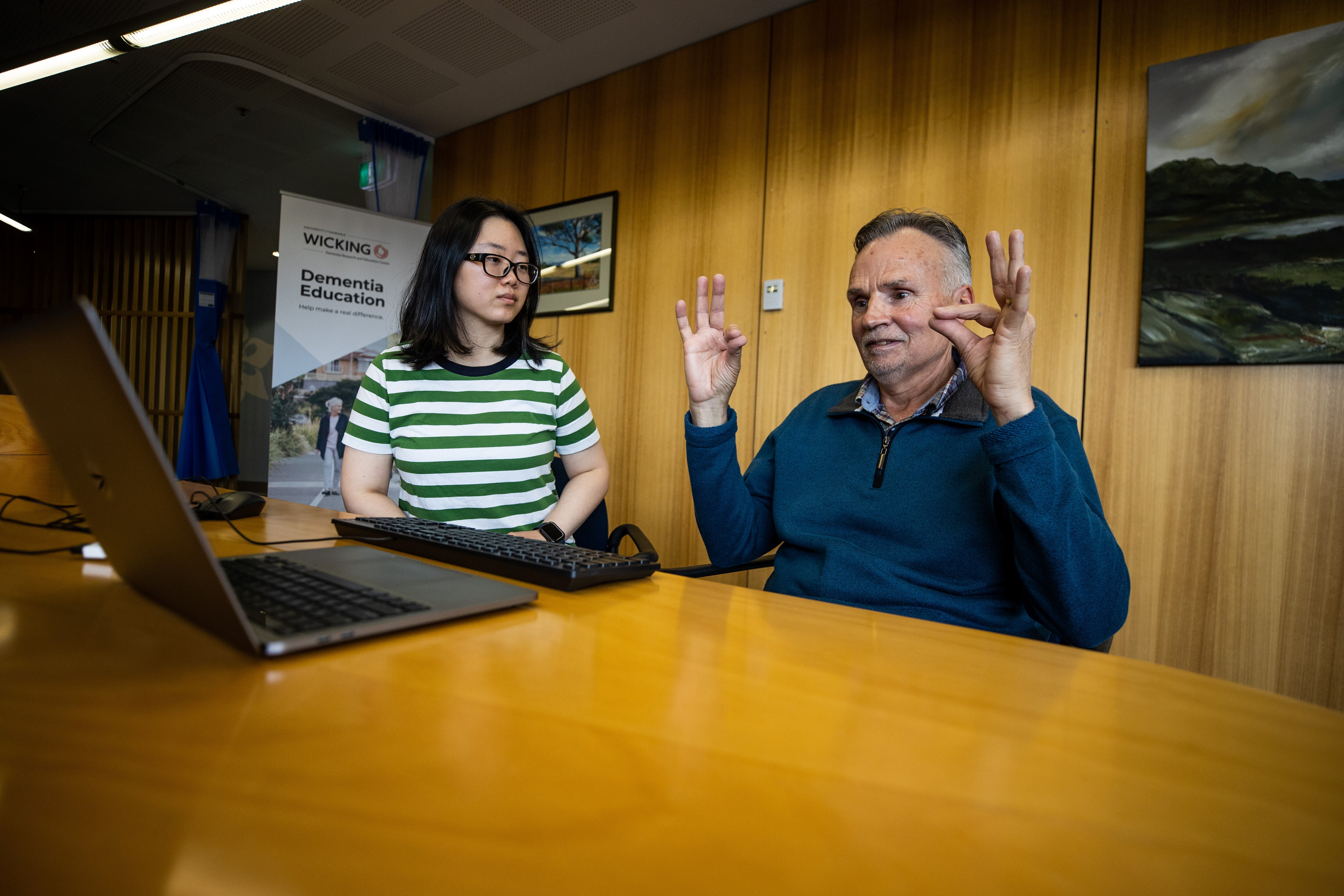 Jon Moore sits at a table in front of a laptop and holds up his hands, a young woman sits beside him watching