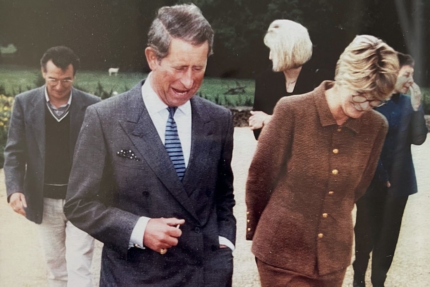a man in a suit and a woman in a brown blazer walk through a country estate's garden