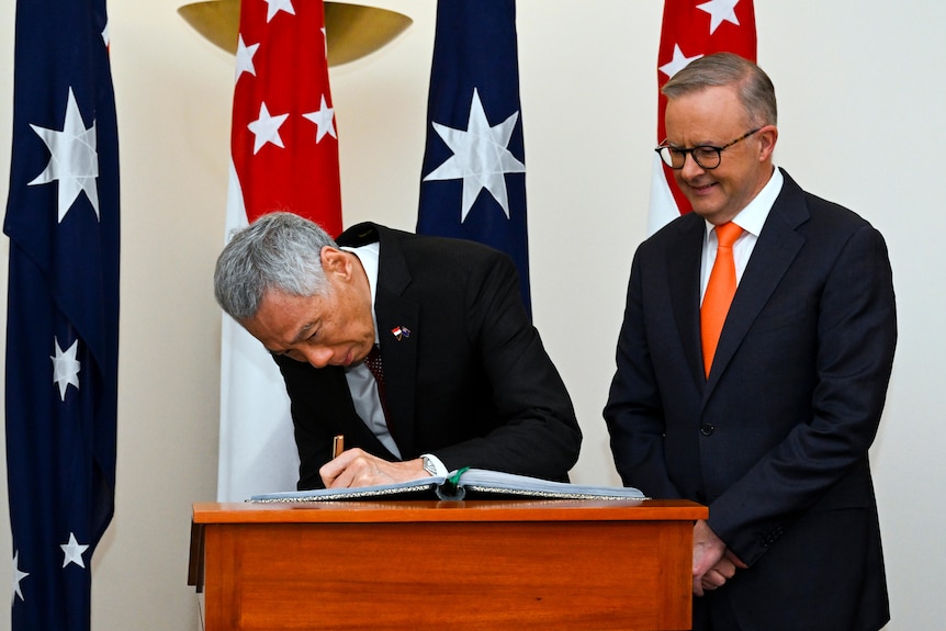 A suited man signs a book while another smiles and watches on, with the flags of Australia and Singapore behind.
