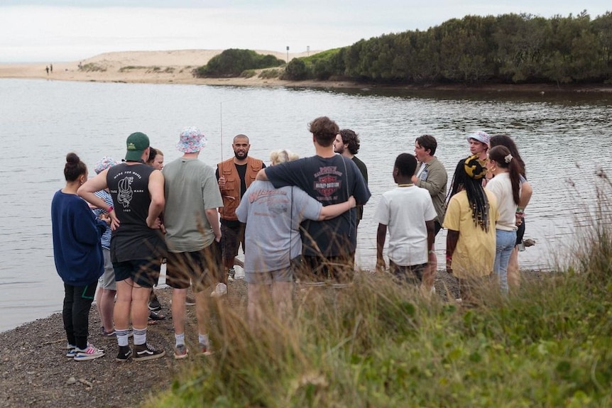 A group stand around a lagoon watching someone hold a fishing rod.