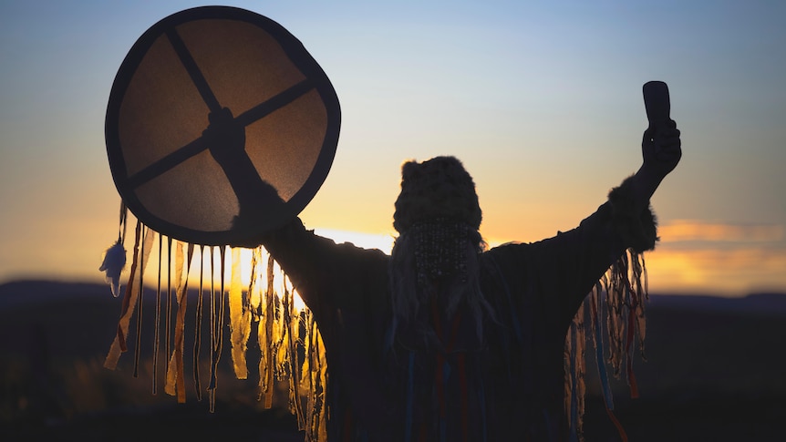 A person holds a shamanic drum in the air at sunrise