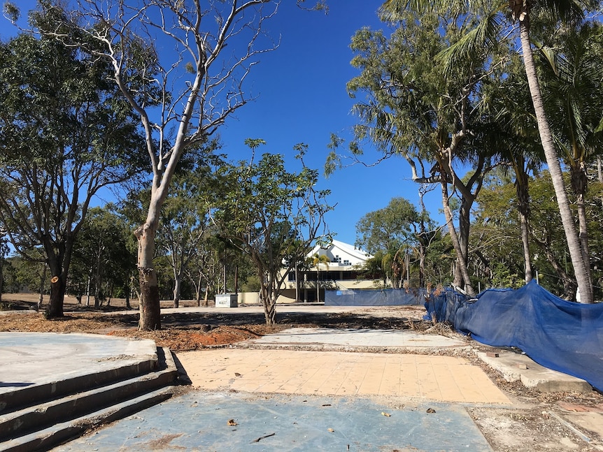 Cement slabs on ground are surround by trees. A building fence looks rundown and leads to an old building.