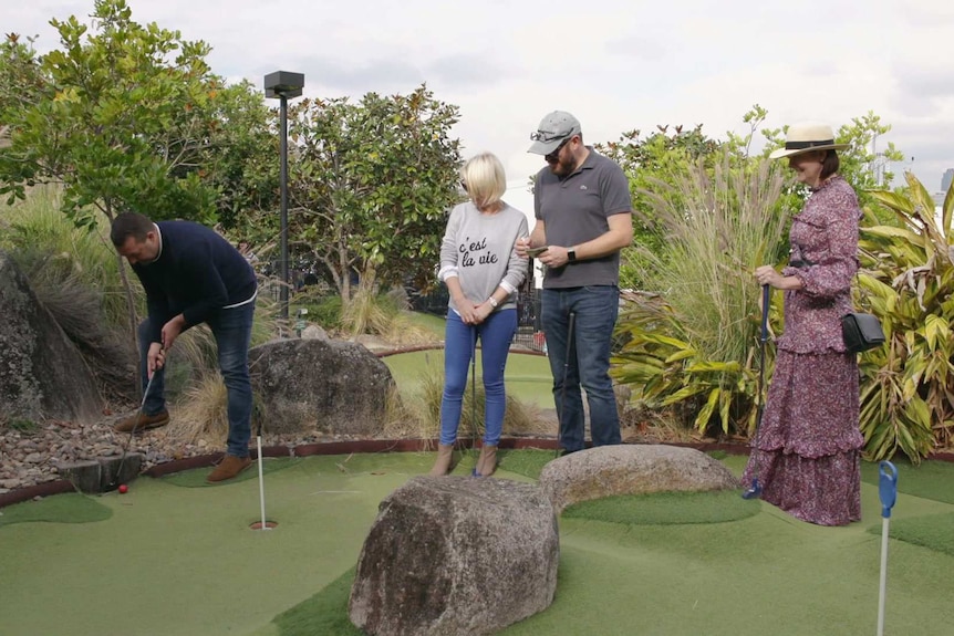 Four people stand on a mini-gol course together. Three watch as one man putts.