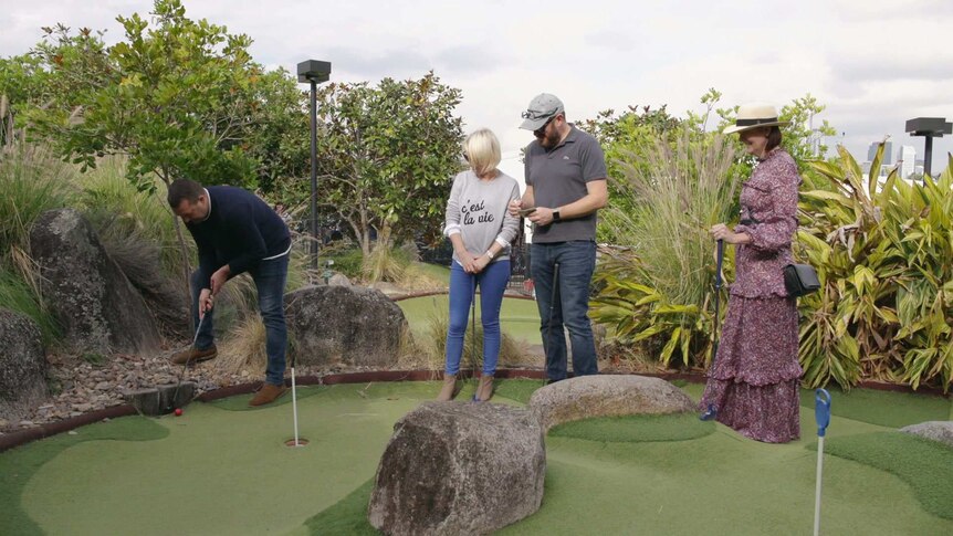 Four people stand on a mini-gol course together. Three watch as one man putts.