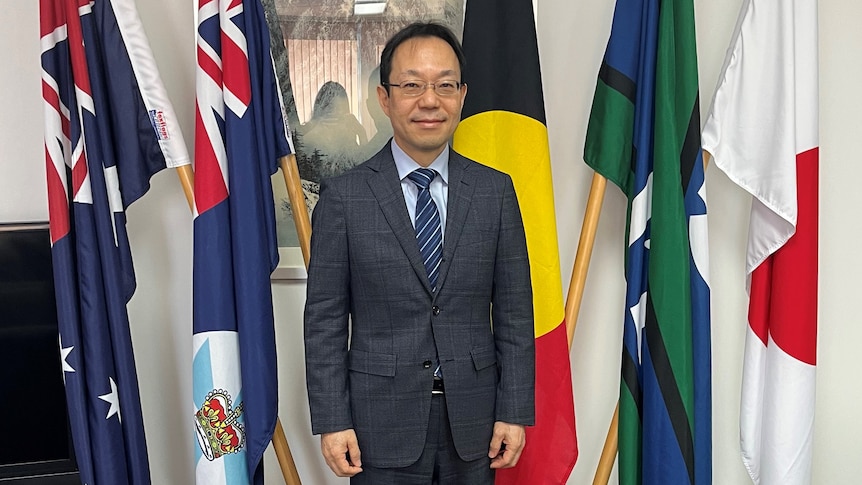 A Japanese man in a suit and tie smiles in front of Australian, Queensland, Aboriginal, Torres Strait and Japanese flags.