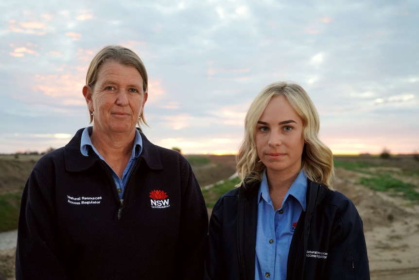 Photo of two women standing in the open, with a paddock behind them. Both are wearing chambrey shirts and badged navy jumpers