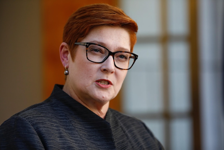 A woman with short red hair with glasses mid-sentence with the background blurred out