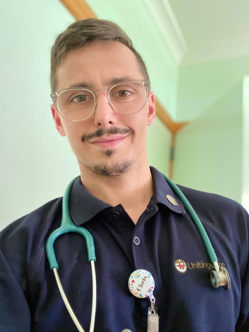 A man wearing nursing scrubs, a stethoscope and glasses.  