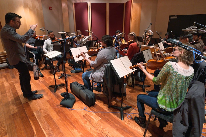 A group of musicians sit in lines poised with a mix of instruments, mainly violins, while a conductor leads them, gesticulating.