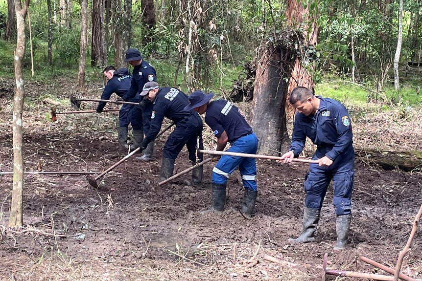 A line of police in blue uniforms use rakes to dig through muddy ground.