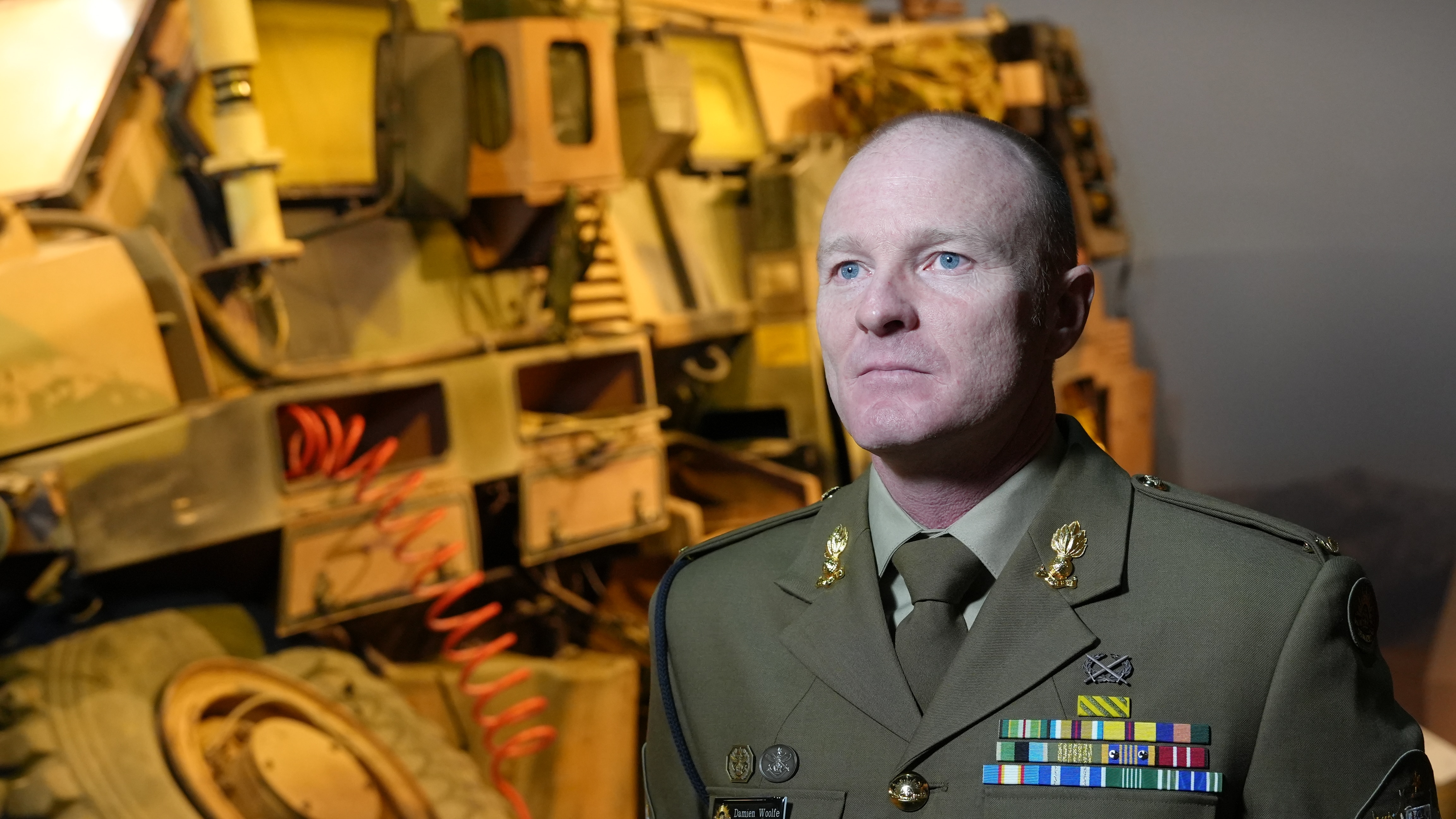 A bald man in military uniform stands under bright lights in front of a military vehicle on display.