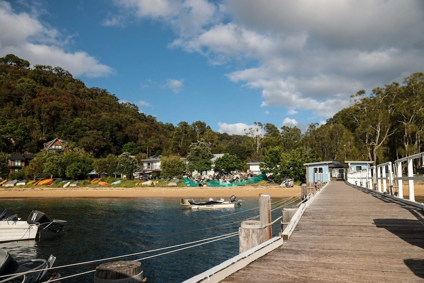 An image of Great Mackerel Beach with a wharf and a boat in the foreground and homes surrounded by trees in the background.