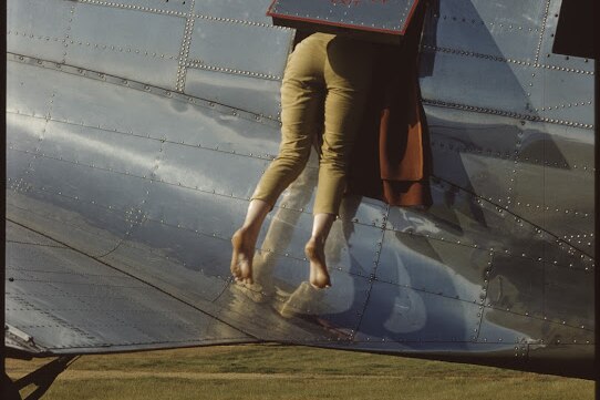 A woman slides backwards out of the hatch on the side of a silver airplane. 