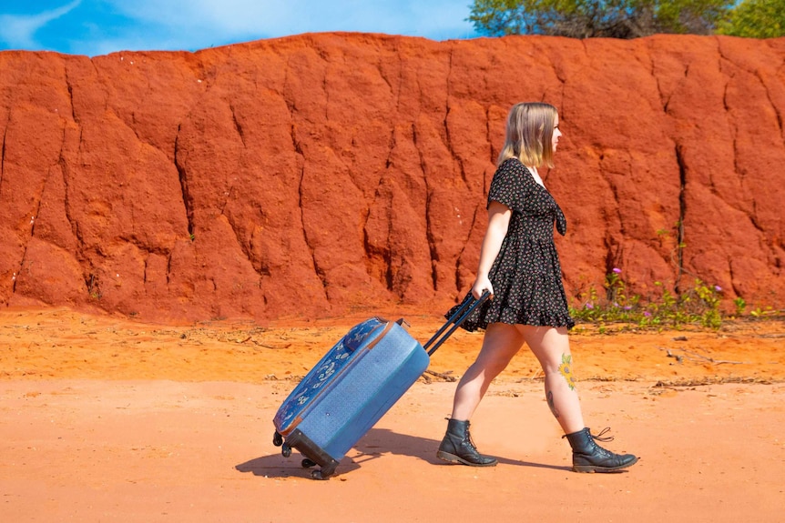 A girl in a summer frock pulls her trolley bag in front of a red cliff face