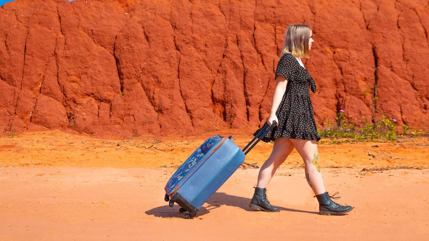 A girl in a summer frock pulls her trolley bag in front of a red cliff face