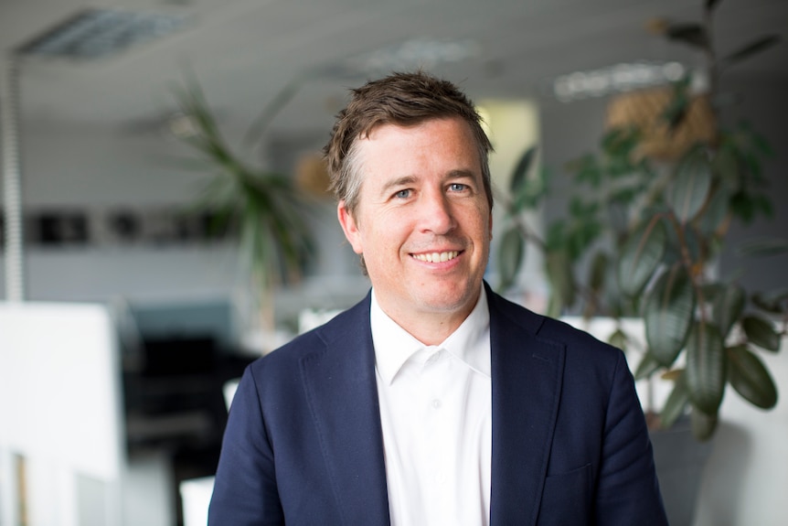 Chris wears a navy suit jacket and white shirt and smiles as he sits in a leafy green office.