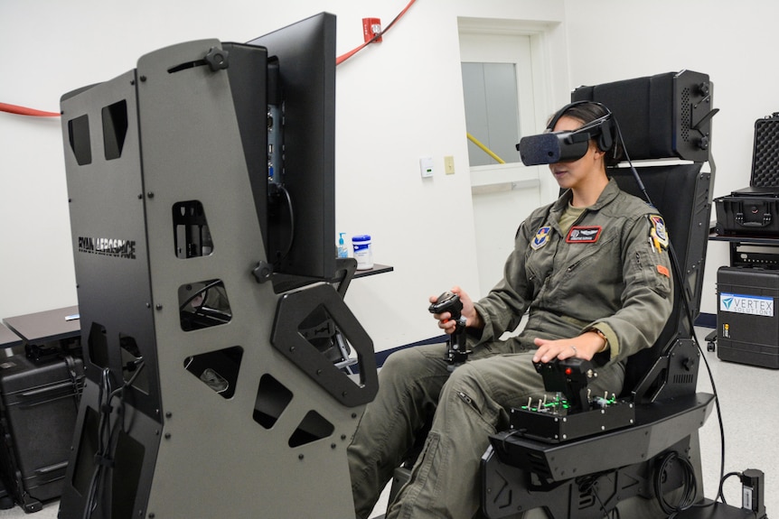 A woman wearing air force fatigues wears virtual reality goggles and sits in a large flight simulator 